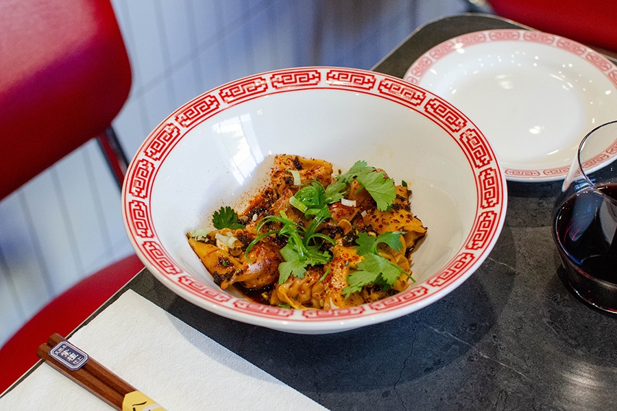 A white bowl with a red border is full of dumplings that are covered in chili flakes and other garnishes.