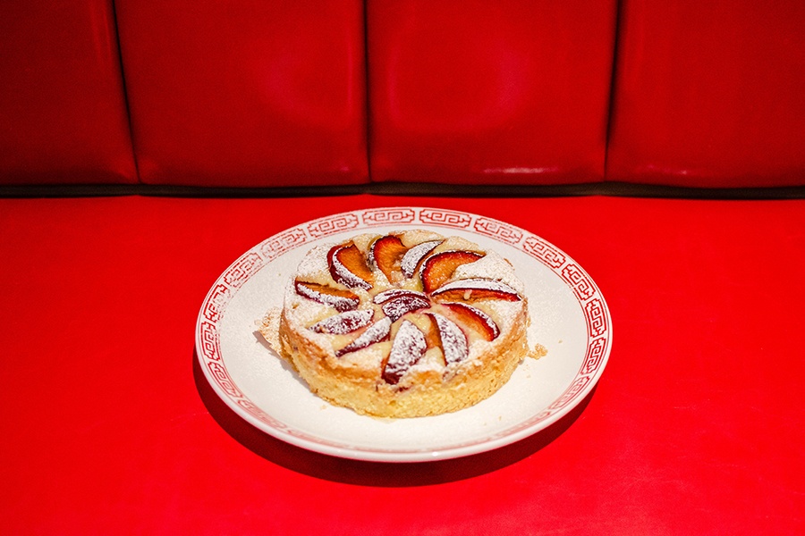 A cake is topped with sliced plums and displayed on a red booth.