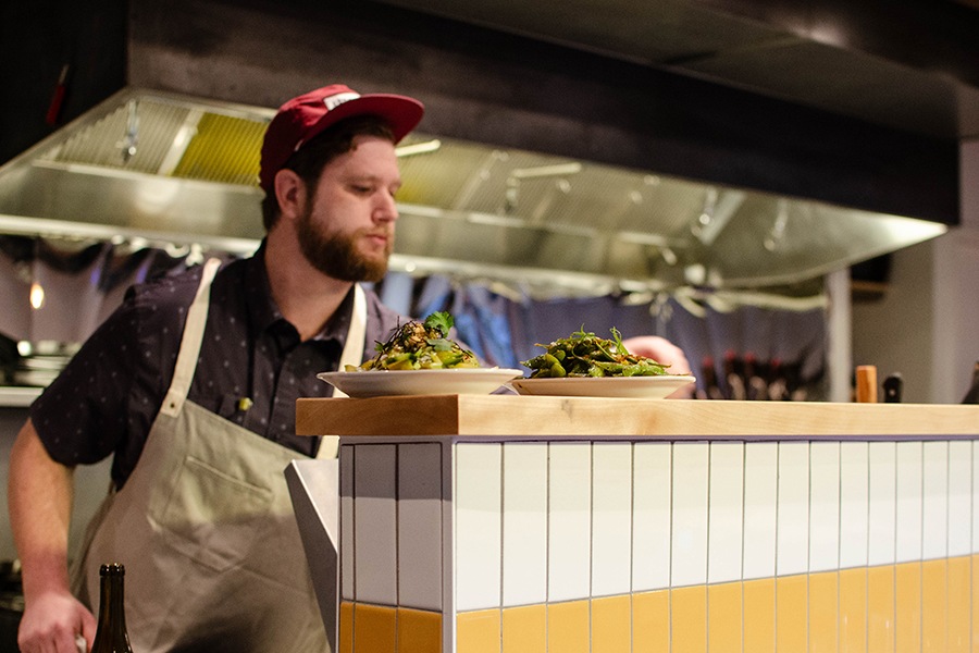 A chef in an apron and baseball cap adjusts a couple vegetable dishes sitting atop the pass in an open kitchen.