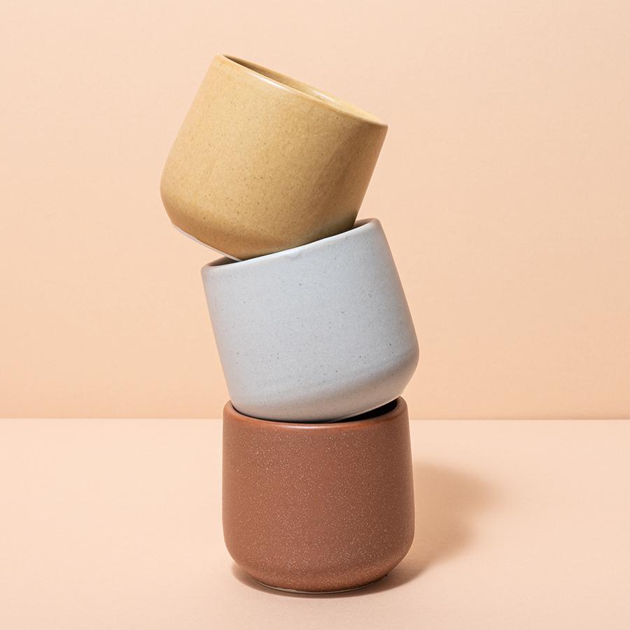 Three small ceramic cups—one brown, one off-white, and one yellow-beige—are stacked atop each other.