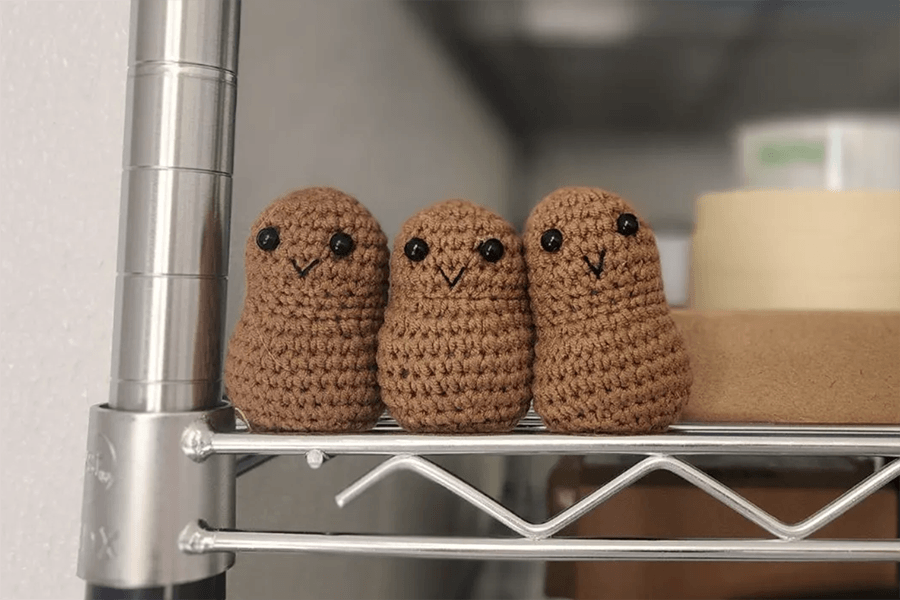 Three small brown crocheted plushes with black beady eyes and embroidered smiles sit on a shelf.