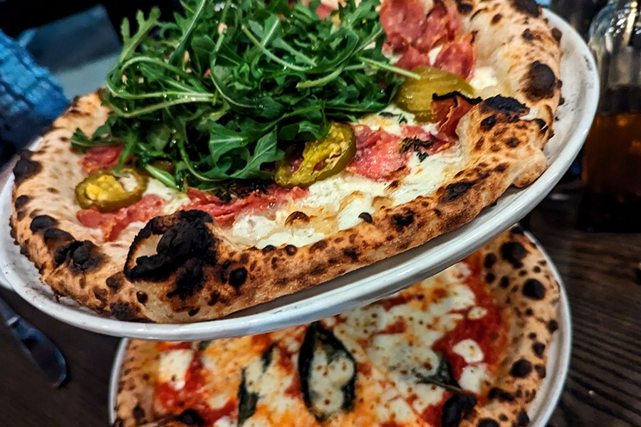 Two wood-fired pizzas with char-spotted crusts are stacked on a holder. The top one is garnished with lots of arugula.