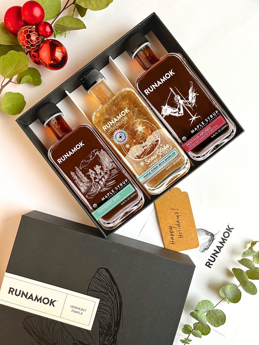 Three different maple syrups are presented in a gift box, surrounded by holiday accoutrements.