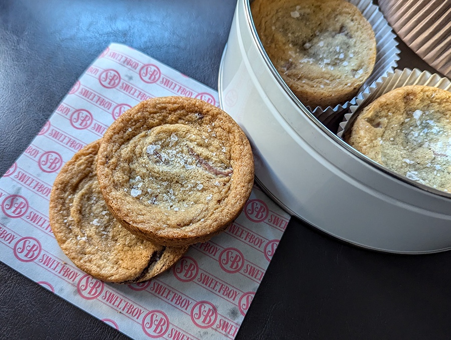 Two big chocolate chip cookies, sprinkled with salt, sit next to a tin with more cookies in it.