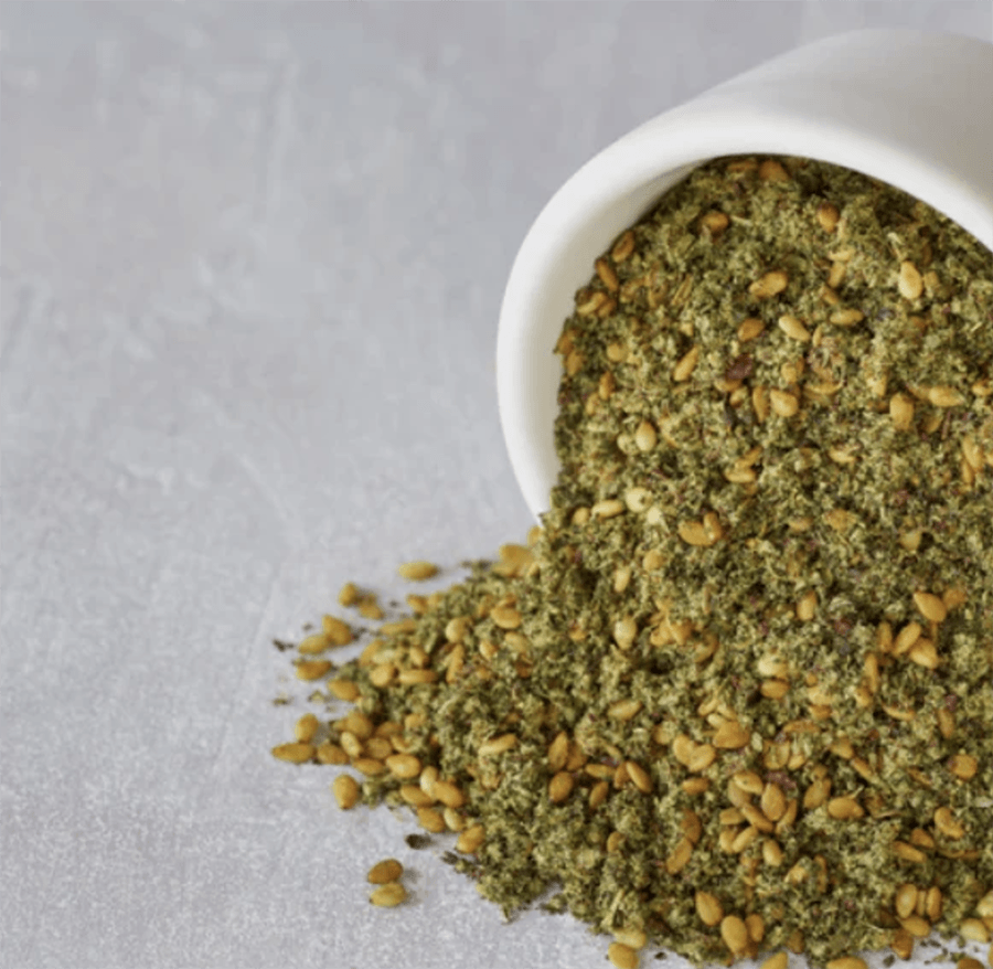 The spice blend za'atar, including dried thyme and sesame seeds, spills out of a white jar.