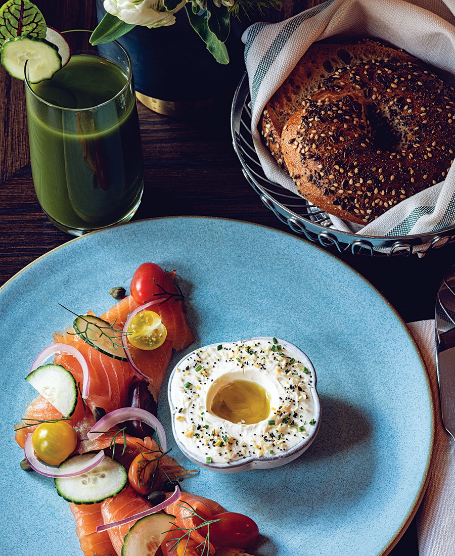 A basket of bagels sits next to a plate with lox, tomatoes, cucumbers, onions, cream cheese, and other accoutrements.