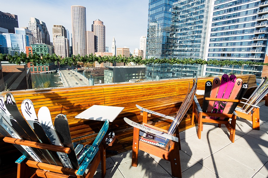 Adirondack chairs built of skis are on a rooftop deck looking out toward the Boston city skyline.