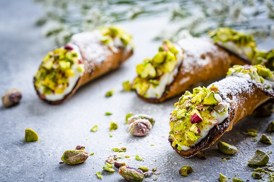 Three pistachio-topped cannoli sit on a powdered sugar-dusted surface.