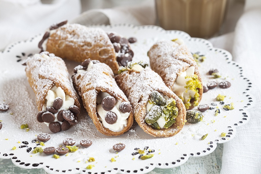 Powdered sugar-dusted cannoli, some with pistachios and some with chocolate chips, are displayed on a plate.