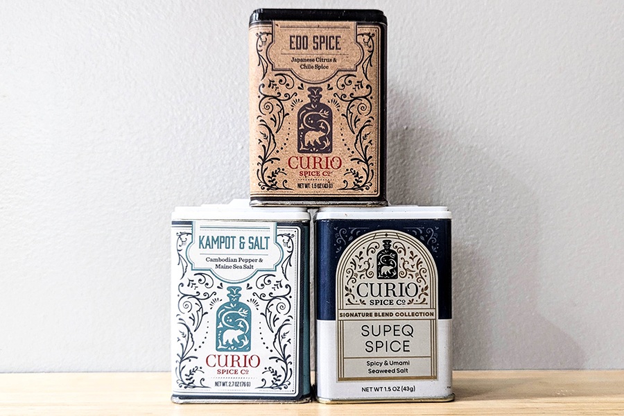 Three tins of different spices are displayed on a wooden counter: kampot and salt, edo spice, and supeq spice from Curio Spice Company.