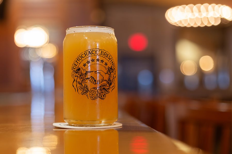 A pint of beer is in a glass with Democracy Brewing branding, including imagery of a handshake over hops.