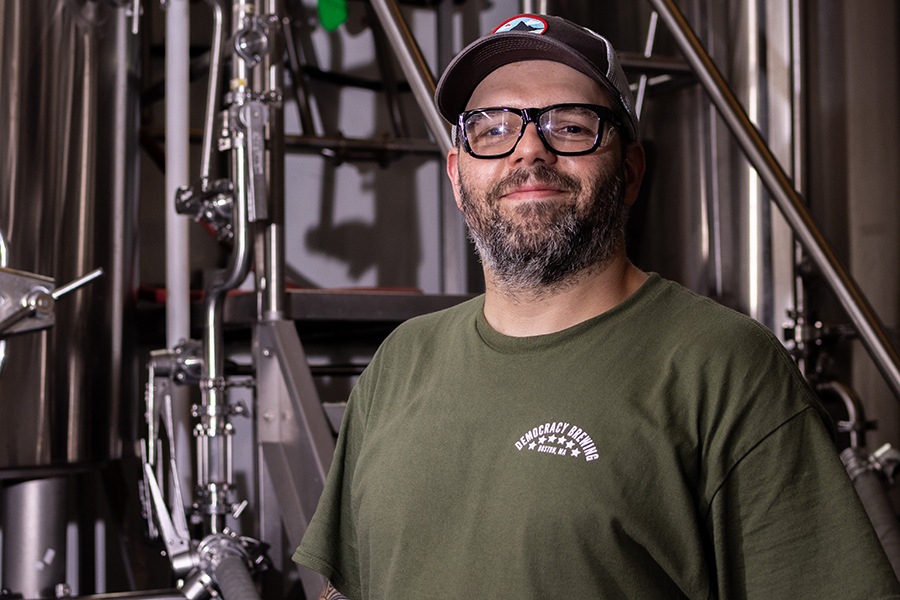 A man with glasses, a beard, and a baseball cap smiles at the camera, standing in front of brewing equipment.