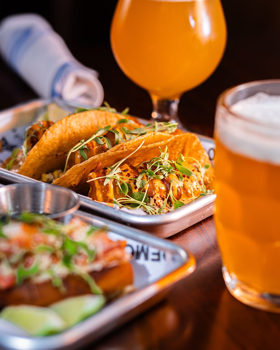 Trays of tacos sit on a table alongside two glasses of beer.