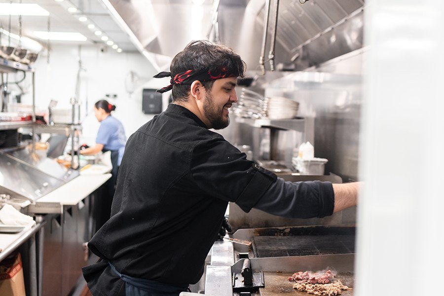 A chef in a restaurant kitchen stands over the stovetop, smiling.