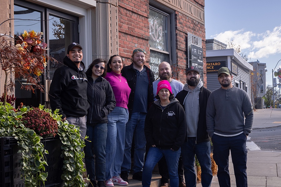 A group of eight people pose, smiling, in front of a red brick building.