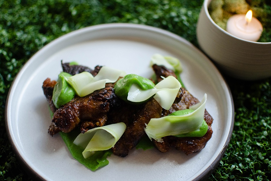 A browned, crispy vegetable is garnished with spots of bright green puree and strips of pale green broccoli stem, with the plate on a backdrop of faux microgreens.