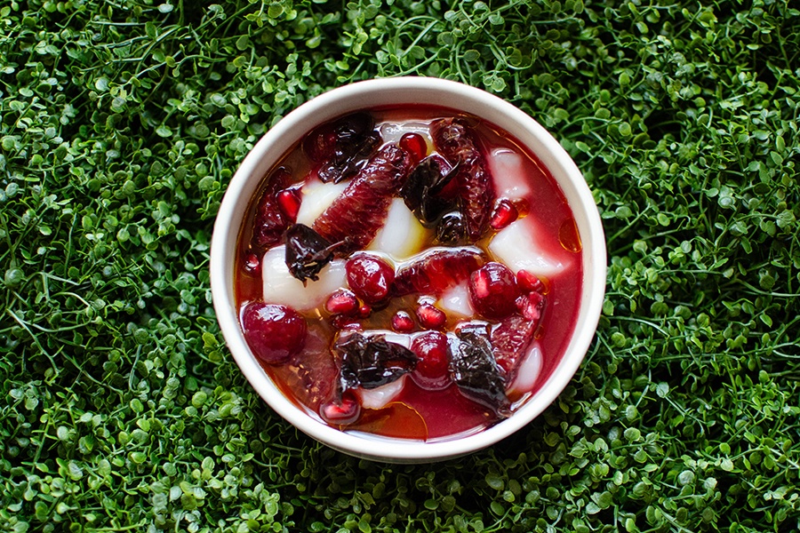 Pieces of raw scallop are presented on a circular plate with bold red ingredients, including blood orange segments and pomegranate seeds. The plate sits on a backdrop of faux microgreens.