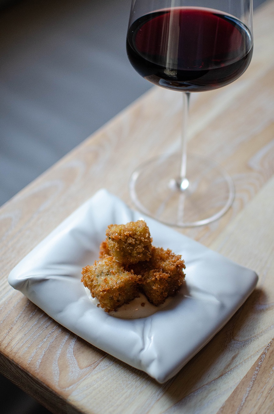 Small, crispy breaded cubes sit atop a plate that looks like a white pillow, with a glass of red wine behind.