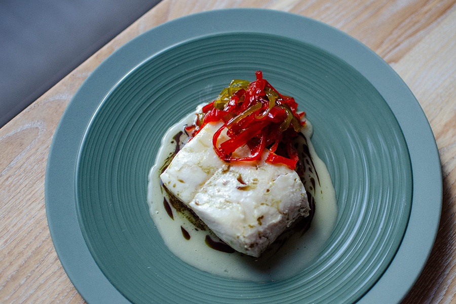 A poached fish filet sits in a white-ish sauce and is garnished with strips of preserved red and green peppers on a pale blue plate.