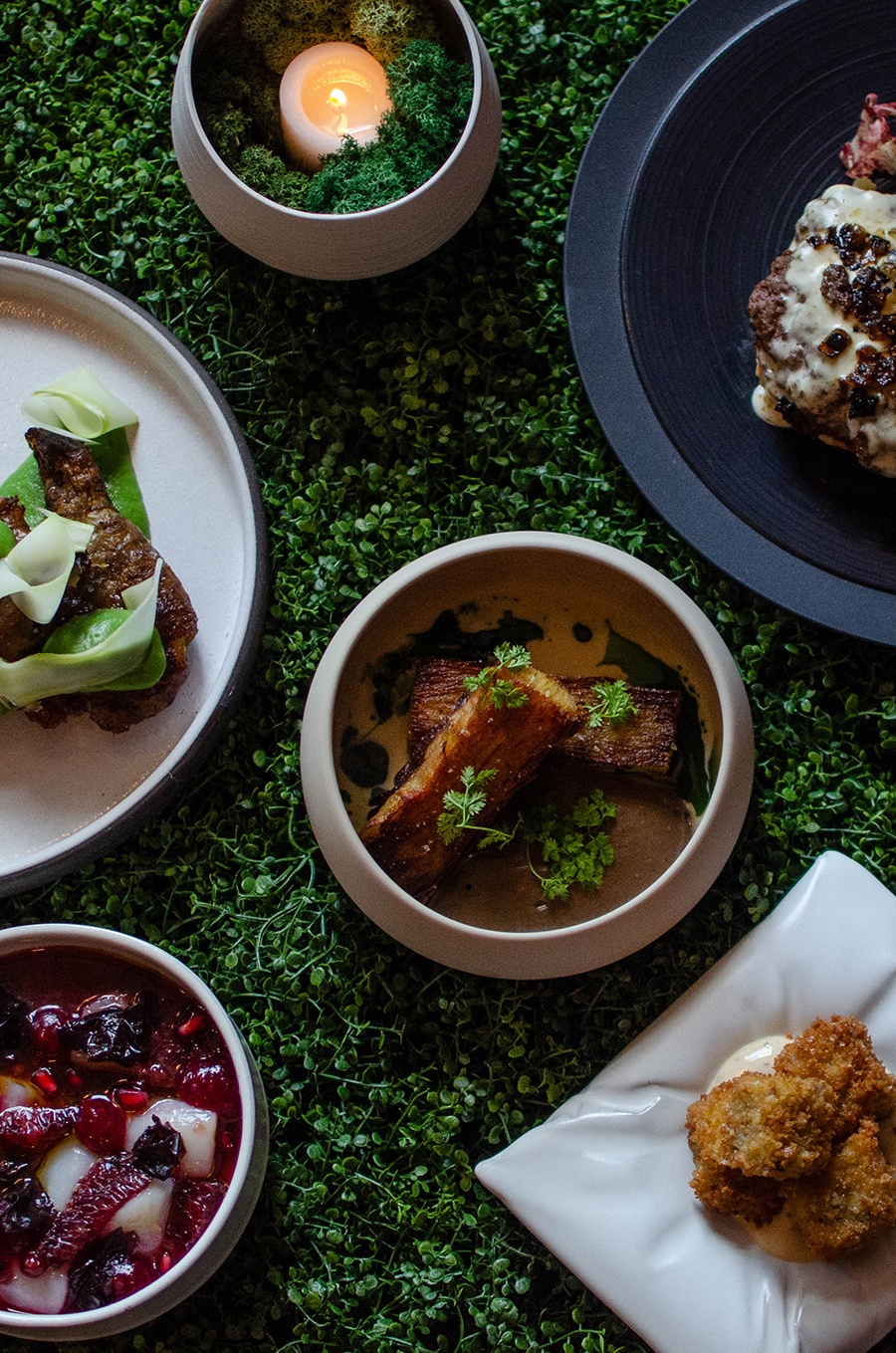 An overhead view shows five food dishes on a faux microgreens backdrop. The dishes include a crudo, crispy potato mille-feuille, and more.