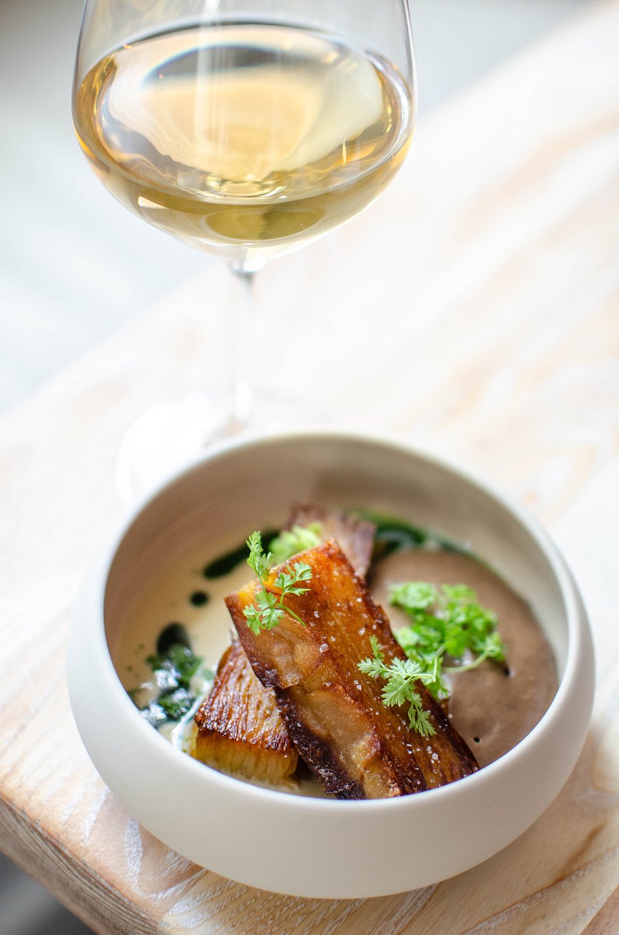 Crispy potato mille-feuille garnished with greens sits in a thick beige sauce in a small bowl, a glass of white wine behind it.