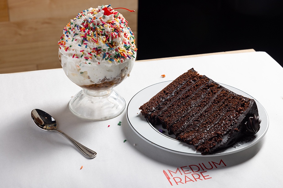 A big ice cream sundae and a slice of chocolate layer cake sit on white paper on a table, stamped in red with Medium Rare.