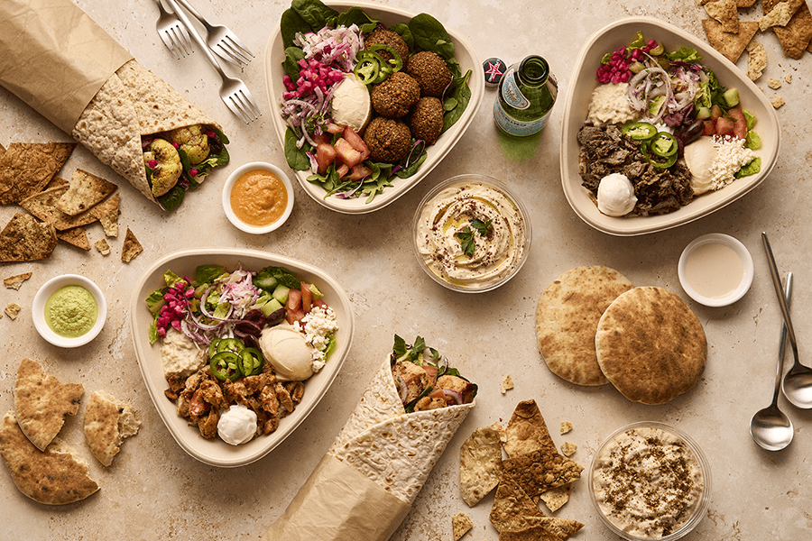 Overhead view of casual Middle Eastern-inspired grain and green bowls on a beige background scattered with bits of pita, sauces, and more.