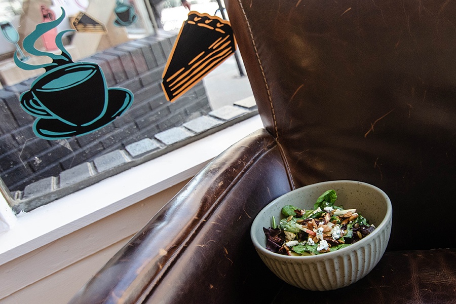 A cheese-, apple-, and nut-topped salad sits in a bowl in a brown leather armchair, next to a window with decals of a coffee cup and pie slice.