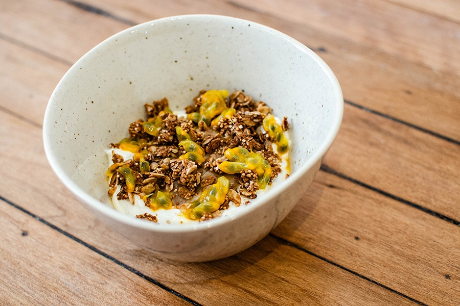 A bowl of white yogurt is topped with. yogurt and passionfruit seeds and sits on a wooden surface.