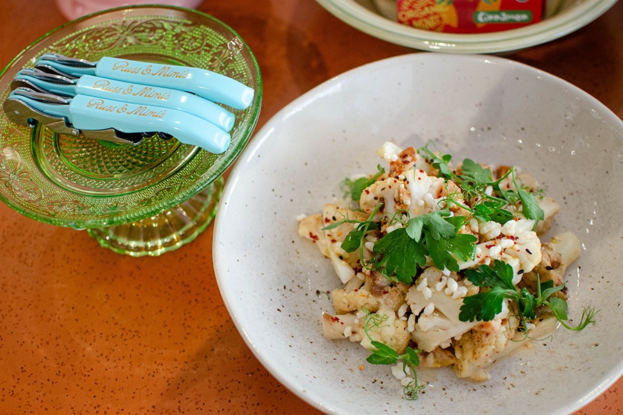 A bowl of roasted cauliflower topped with greens and crispy rice sits next to Russ & Mimi's branded light blue bottle openers.