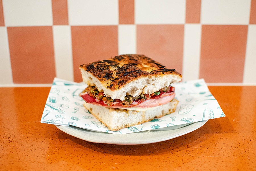 A sandwich with deli meats and cheese piled on focaccia sits on a plate on an orange countertop.