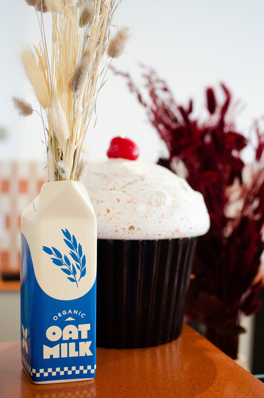 A vase with plants in it looks like an oat milk container, and a giant cupcake sculpture sits behind it on an orange counter.