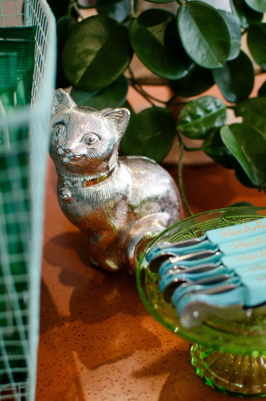 A silver cat figurine, greens, and pale blue bottle openers sit on an orange counter.