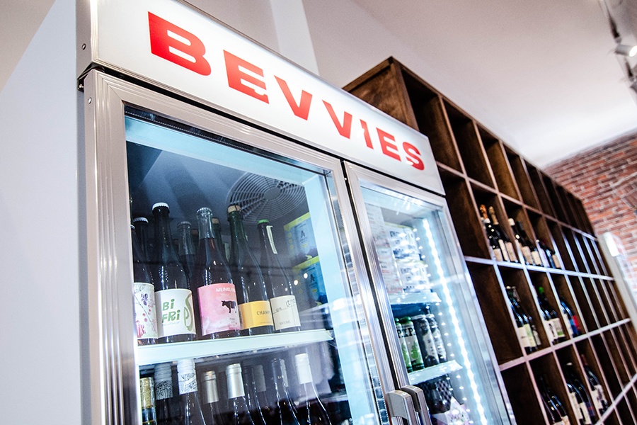 Wine and beer are stocked in a fridge that says "Bevvies" in big red letters, with wooden shelves of wine bottles visible in the background.