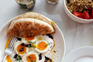 Overhead view of eggs in a pool of labneh and olive oil with a pita, plus a yogurt parfait in a nearby bowl.