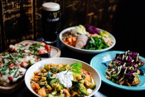 Four colorful vegetarian dishes are spread on a table next to a pint glass of Guinness.