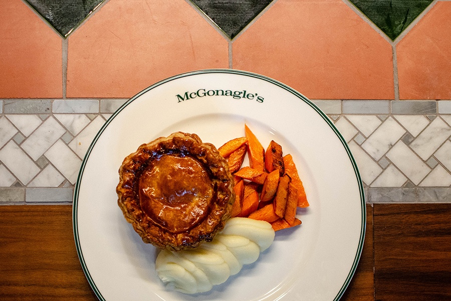 A shiny, browned mini pie is accompanied by roasted carrots and creamy mashed potatoes, served on a plate that says McGonagle's.