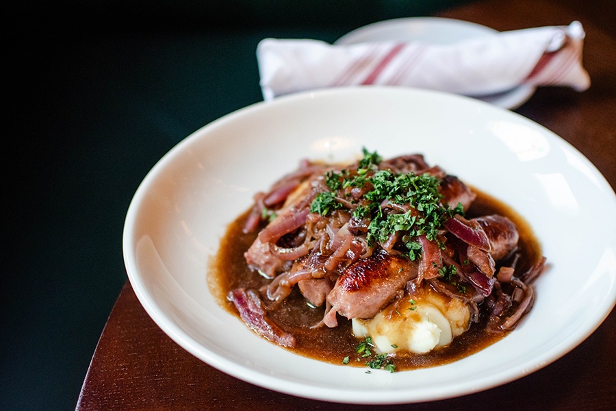 Sausages topped with herbs and caramelized onions sit atop mashed potatoes, all smothered in a thin brown gravy.