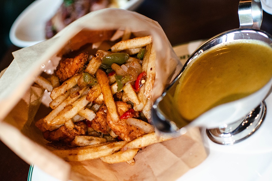 A paper bag is opened to reveal fries, browned chicken bits, and bell pepper strips next to a gravy boat of curry.