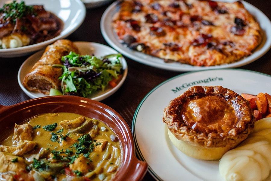 A spread of food at a modern Irish pub, including a small round pie with mashed potatoes, diced chicken in a yellow-brown curry, a pepperoni pizza, and more.