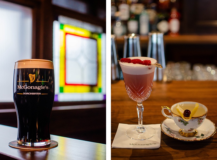 A pint of dark beer in a glass that says McGonagle's Dorchester sits in front of stained glass. A pink cocktail topped with raspberries and a cocktail in a floral tea cup sit on a wooden bar.