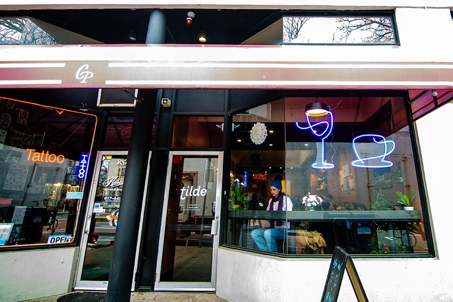 The exterior of a coffeeshop reads Tilde, and it has blue neon signage in the window in the shape of a wine glass and a coffee cup.