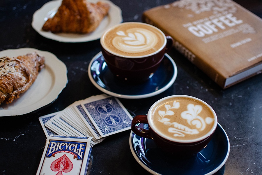 Lattes, pastries, a book about coffee, and playing cards are displayed on a table.