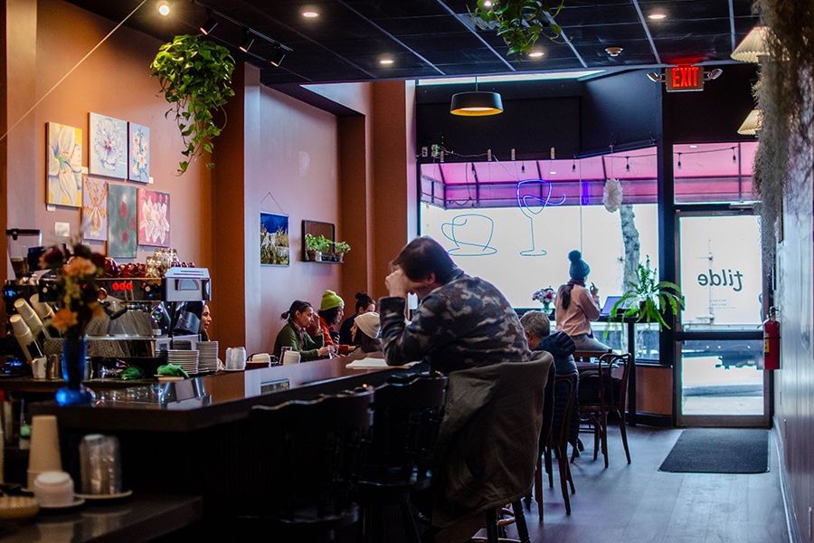 A small cafe decorated with pops of greenery, floral paintings, and neon signage of a coffee cup and a wine glass is partially filled with customers.