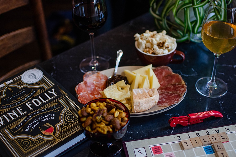 A table is spread with a small charcuterie plate, bowls of nuts and popcorn, wine, a book about wine, and a scrabble board.