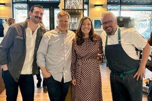 Four smiling people stand in a line in a restaurant, looking at the camera. One is in a chef's apron.