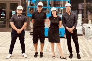 Four people dressed in black clothes and white hard hats smile, lined up in front of an under-construction space in a tall building.