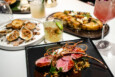 A rack of lamb, focaccia, and cooked oysters are displayed on a white marble surface alongside several cocktails.