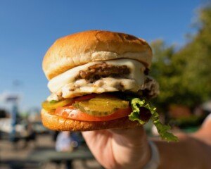 A hand holds up a cheeseburger outdoors.