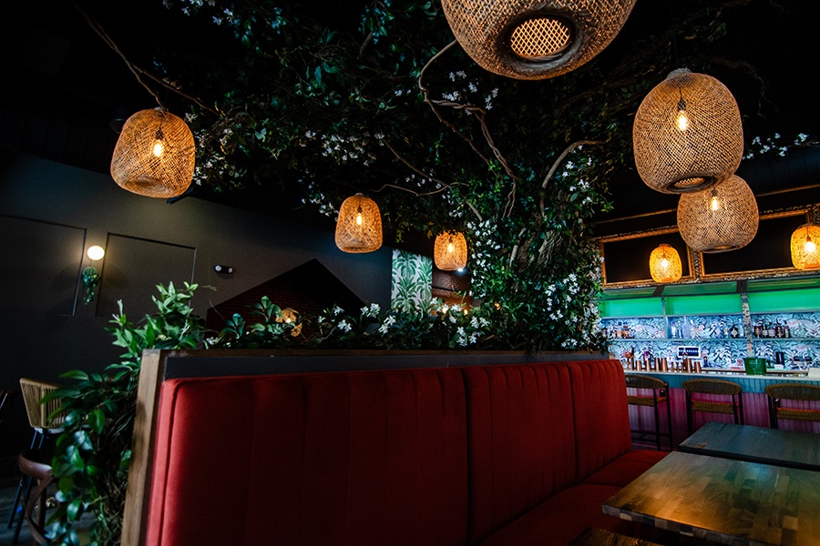 Interior of a restaurant with a big faux greenery-covered tree, from which basket-like lanterns dangle. There's a plush red banquette and several tropical wallpaper patterns.